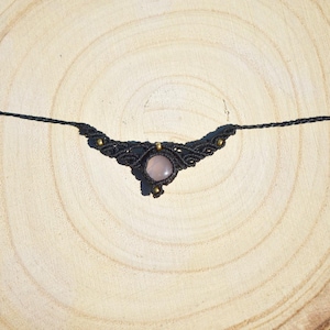 May include: A black macrame necklace with a central rose quartz stone. The necklace features intricate knotting and small gold-colored beads. The necklace is displayed on a light brown wooden surface.