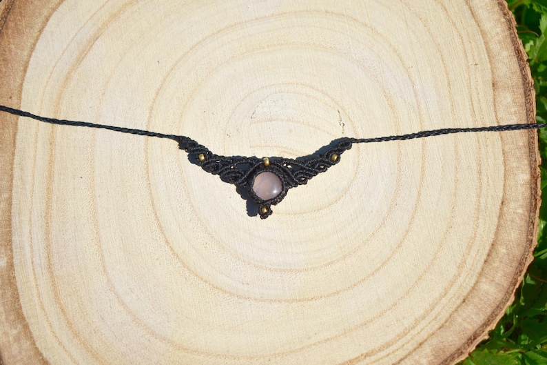 May include: A black macrame necklace with a central rose quartz stone. The necklace features intricate knotting and small gold-colored beads. The necklace is displayed on a light brown wooden surface.