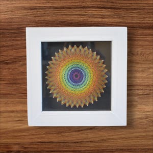May include: A framed string art piece featuring a vibrant rainbow-colored design. The artwork is composed of concentric circles of thread, creating a sunburst pattern. The piece is set in a square white frame, displayed against a wooden background.