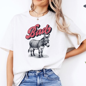 May include: White t-shirt with a graphic of a donkey and the word "Bad" in red script. The donkey is in black and white, standing on a patch of grass. The shirt is a casual style.