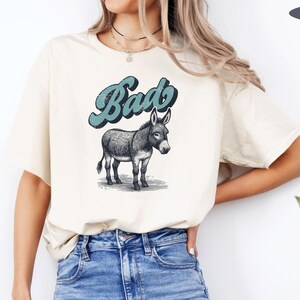May include: A beige t-shirt with a graphic of a donkey and the word "Bad" in blue text. The model is wearing the shirt with blue jeans.