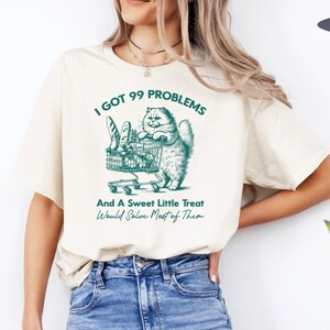 May include: A beige t-shirt with a green graphic of a cat pushing a shopping cart full of bread. The text "I got 99 problems and a sweet little treat would solve most of them" is printed on the shirt.