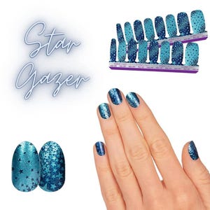 Op de afbeelding: Een set Star Gazer nagellakstrips met een blauwe gradi&euml;ntachtergrond en een donkerblauw sterrenpatroon. De strips worden getoond op twee rechthoekige kaarten en aangebracht op vingernagels, met een glinsterende afwerking en sterrenmotieven.
