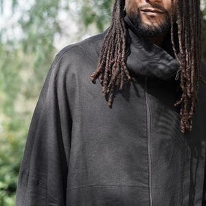 May include: A black jacket with a high collar and a front zip. The jacket features a large pocket on the front and is made of a soft, flowing fabric. The person in the image has long dreadlocks.