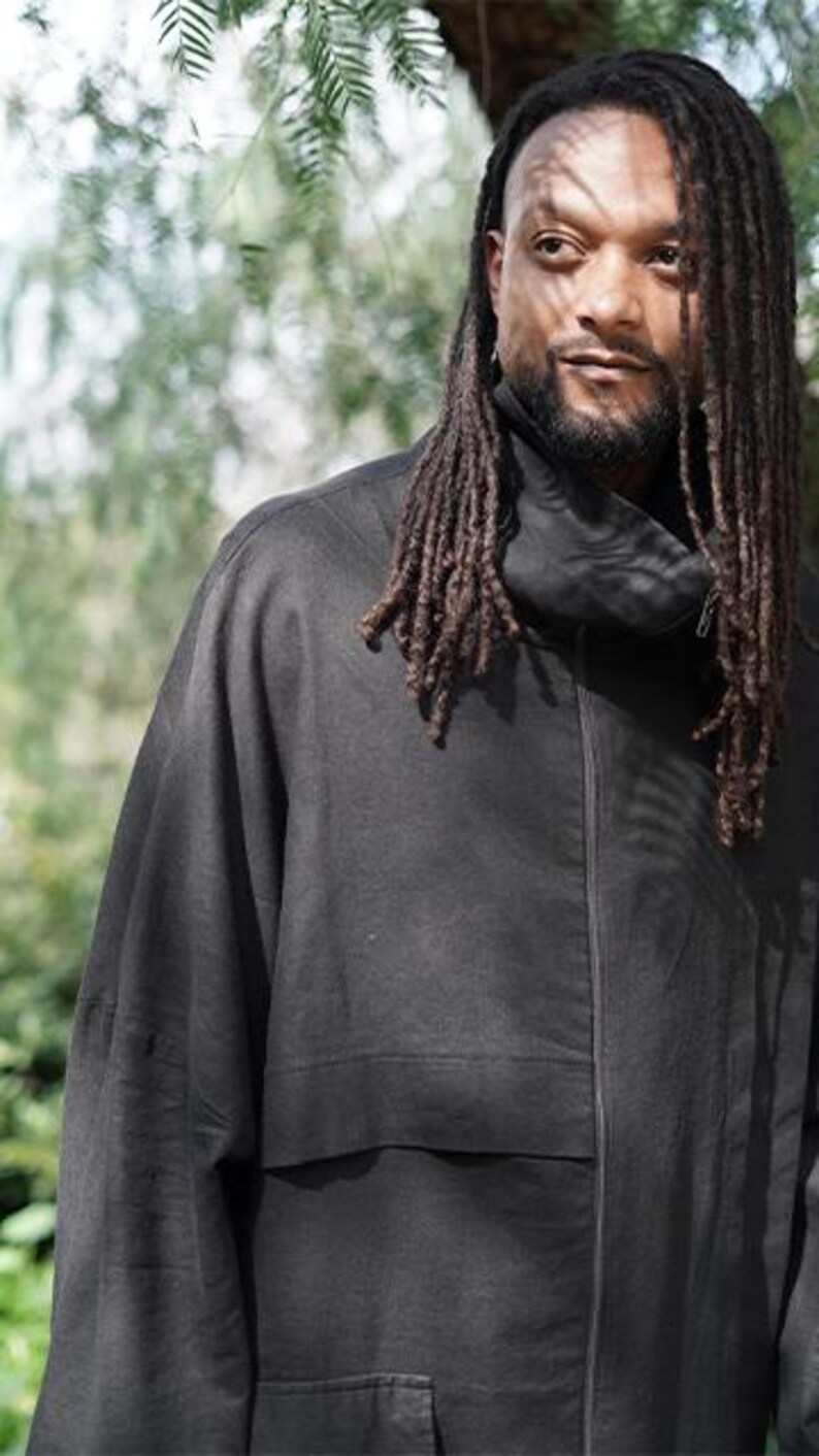May include: A black jacket with a high collar and a front zip. The jacket features a large pocket on the front and is made of a soft, flowing fabric. The person in the image has long dreadlocks.
