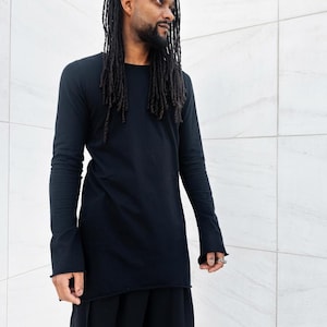 Könnte beinhalten: Ein schwarzes Langarmshirt mit Rundhalsausschnitt und ausgestellten Ärmeln. Das Shirt wird mit einer schwarzen Hose kombiniert. Das Model hat lange Dreadlocks und einen Bart. Der Hintergrund ist eine weiße geflieste Wand.