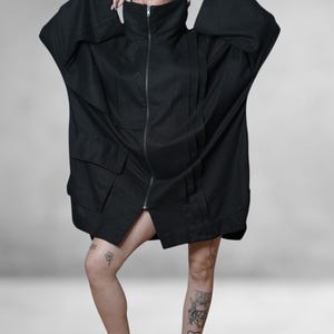 May include: Black oversized jacket with a high collar and a full-length zip. The jacket features large pockets and wide sleeves. The model is wearing the jacket. The jacket is made of a dark fabric.