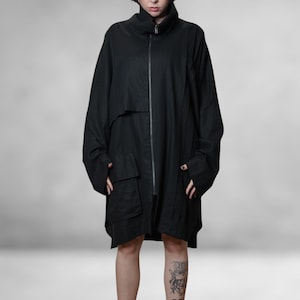May include: A black, oversized coat with a high collar and a full-length zip. The coat has two large front pockets and long sleeves. The model is standing in front of a grey background.