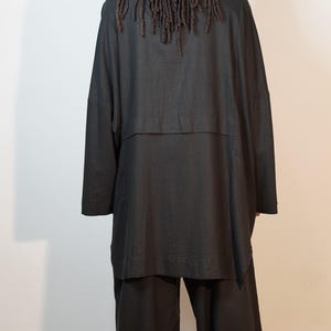 May include: A black outfit, comprising a long-sleeved top and wide-leg trousers. The top features a layered design. The person has long, dark dreadlocks. The outfit is made of a soft, flowing fabric.