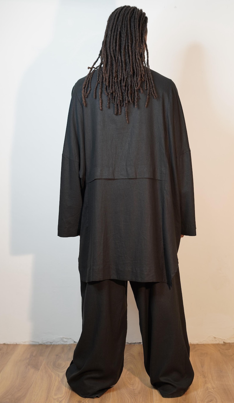 May include: A black outfit, comprising a long-sleeved top and wide-leg trousers. The top features a layered design. The person has long, dark dreadlocks. The outfit is made of a soft, flowing fabric.