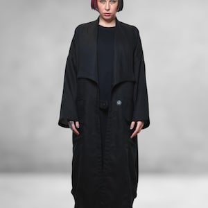 Op de afbeelding: Een lange, zwarte jas met een brede revers en een enkele knoopsluiting. De jas heeft twee voorzakken en wordt over een zwarte top gedragen. Het model draagt zwarte sandalen. De achtergrond is effen grijs.