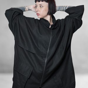 May include: Black oversized jacket with a zip-up front, two large front pockets, and a high collar. The jacket has a relaxed fit and long sleeves. The model is wearing the jacket with their arms behind their head.