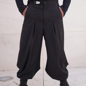 May include: Black wide-leg trousers with a high waist and a silver buckle belt. The trousers have pleats in the front and are paired with black boots. The model is wearing a black long-sleeved top.