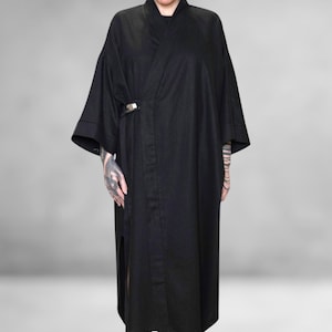 Op de afbeelding: Een lange, zwarte kimono-achtige jurk met wijde mouwen en een wikkel sluiting, vastgezet met een zilverkleurige sluiting. De jurk heeft een hoge kraag en een zijsplit. De persoon draagt zwarte sandalen.