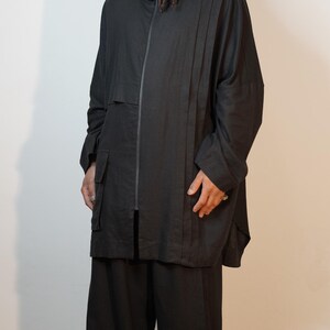May include: A black outfit comprising a zip-up jacket with a high collar, wide-leg trousers, and black shoes. The jacket features a minimalist design with a vertical seam detail. The outfit is made from a lightweight fabric.