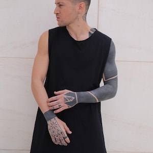 Può includere: Un top nero senza maniche con un orlo lungo e asimmetrico. Il modello ha tatuaggi su braccia e mani. Lo sfondo è un muro liscio di colore chiaro. Lo stile generale è minimalista e moderno.
