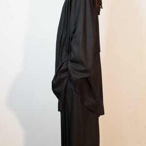 May include: A person in a black outfit, comprising a long coat and wide-leg trousers. The coat features a high collar, and the trousers are floor-length. The person is wearing black shoes. The outfit is made of a dark material.