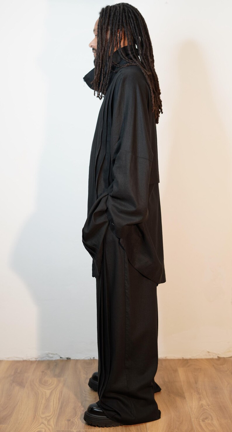 May include: A person in a black outfit, comprising a long coat and wide-leg trousers. The coat features a high collar, and the trousers are floor-length. The person is wearing black shoes. The outfit is made of a dark material.