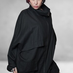 May include: A black, oversized jacket with a high collar and a front zip. The jacket features large pockets and a unique, flowing design. The model has red and black hair.
