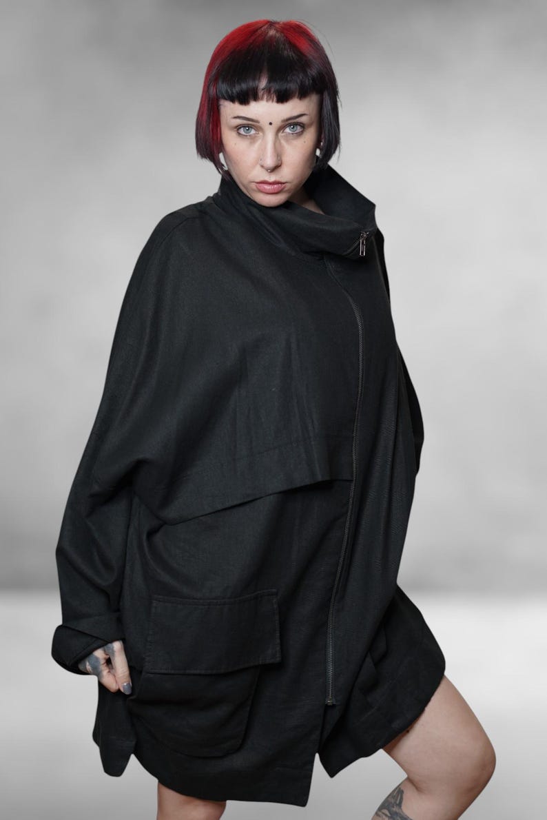 May include: A black, oversized jacket with a high collar and a front zip. The jacket features large pockets and a unique, flowing design. The model has red and black hair.
