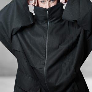 May include: A black jacket featuring a high, zipped collar and oversized sleeves. The jacket's design is minimalist, with vertical seams adding detail. The person's face is partially hidden by the collar. The fabric appears to be lightweight.
