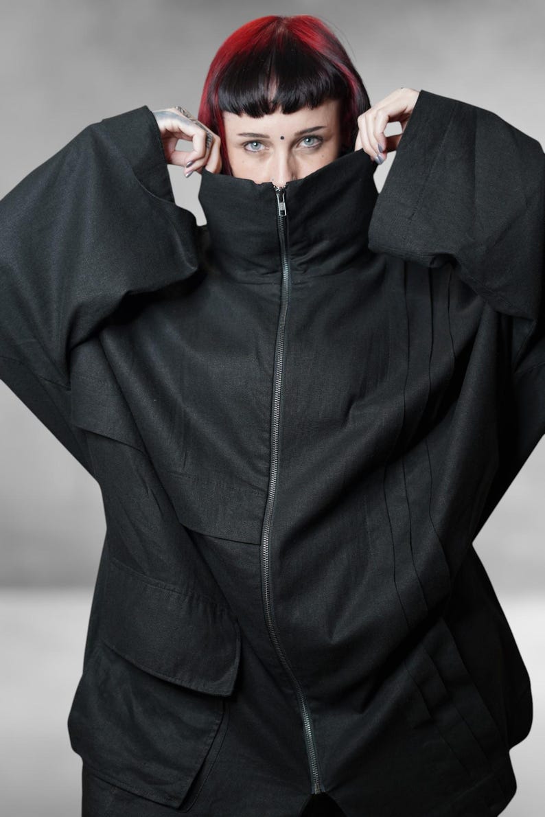 May include: A black jacket featuring a high, zipped collar and oversized sleeves. The jacket's design is minimalist, with vertical seams adding detail. The person's face is partially hidden by the collar. The fabric appears to be lightweight.
