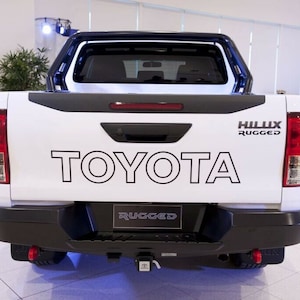May include: White Toyota Hilux Rugged pickup truck with a black roll bar and black bumper. The truck has a black tailgate and a black rear bumper. The truck has a black tow hitch and a black number plate holder. The truck has a black spare tyre cover and a black rear window.