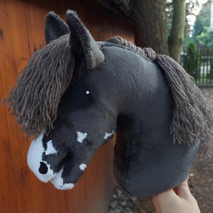Puede incluir: Una cabeza de caballo de peluche gris con una larga melena gris y marcas blancas en el hocico. El caballo tiene un ojo negro y está sujeto por una mano. El juguete está diseñado para ser sostenido como un caballo de palo.