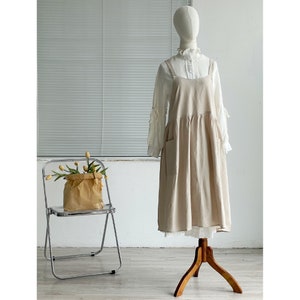 Peut inclure: Une robe en lin beige avec une bordure en dentelle blanche et un chemisier blanc en dessous. La robe a une jupe pliss&eacute;e et un design simple.