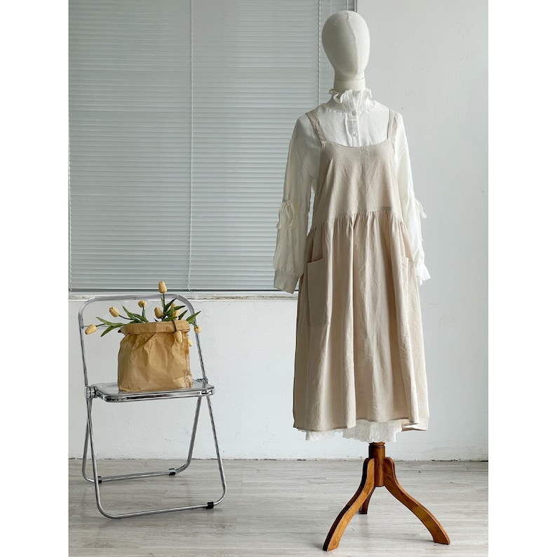 Peut inclure: Une robe en lin beige avec une bordure en dentelle blanche et un chemisier blanc en dessous. La robe a une jupe pliss&eacute;e et un design simple.