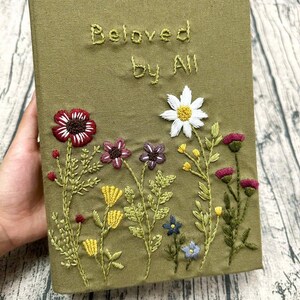 Op de afbeelding: Olijfgroene notitieboekomslag met geborduurd bloemmotief. De omslag heeft de tekst "Beloved by All" in een gestikt schrift. Het bloemenborduurwerk omvat een rode bloem, een witte madelief en andere kleurrijke bloemen.