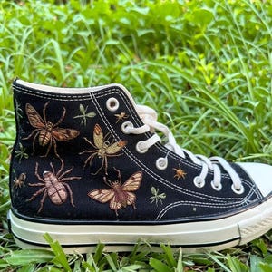 Könnte beinhalten: Schwarzer High-Top-Sneaker mit gestickten Insekten, darunter Spinnen, Motten und Bienen. Der Schuh hat weiße Schnürsenkel, eine weiße Sohle und ein schwarzes Canvas-Obermaterial. Die Insekten sind in Braun-, Beige- und Grüntönen gehalten.
