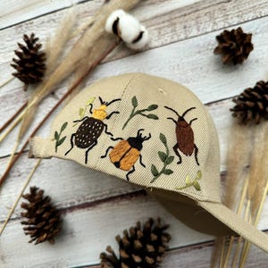 May include: A beige baseball cap with embroidered insects and leaves. The insects are brown, orange, and yellow.