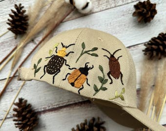 May include: A beige baseball cap with embroidered insects and leaves. The insects are brown, orange, and yellow.