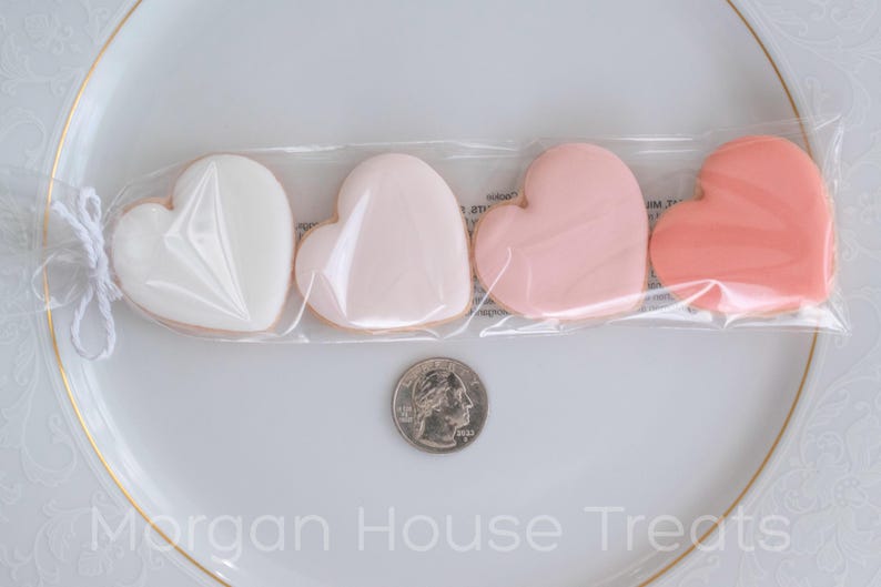 Single sleeve of four pink ombre cookies resting on white china plate with quarter for size reference. Ingredient label on back of sleeve slightly visible on white plate background. Morgan House Treats watermark at bottom of image.