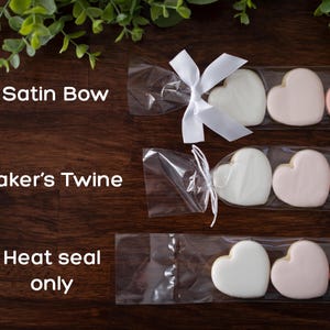 Cookie sleeves laying on wooden table showing the different closure options. Cookie sleeves can be tied closed with a white satin ribbon bow, white baker's twine, and heat seal only (no tie).