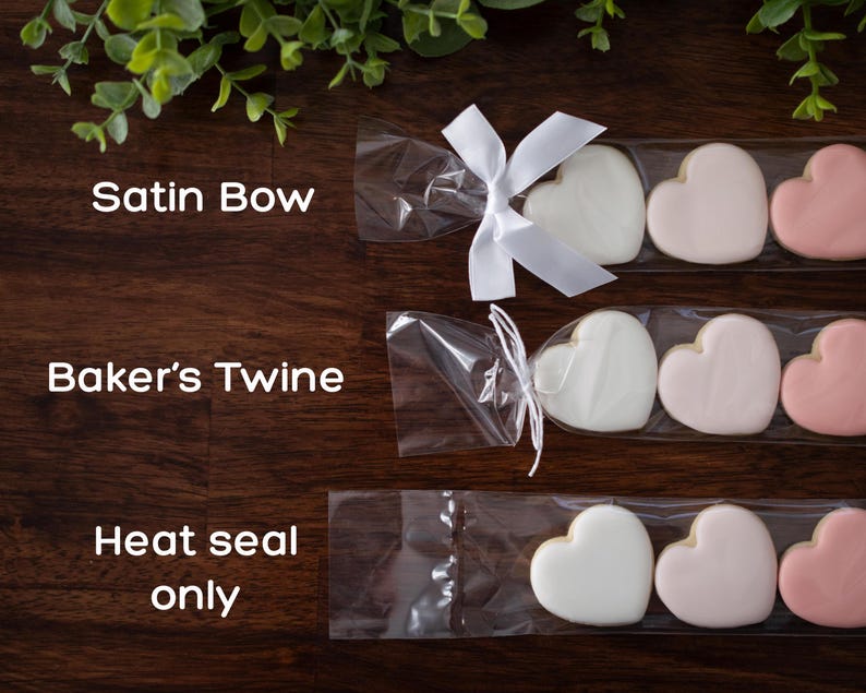 Cookie sleeves laying on wooden table showing the different closure options. Cookie sleeves can be tied closed with a white satin ribbon bow, white baker's twine, and heat seal only (no tie).