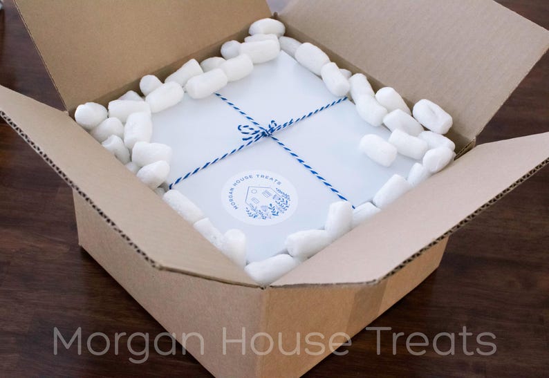 White bakery box packed in open cardboard shipping box showing white compostable packing peanuts. Morgan House Treats watermark on bottom of photo.