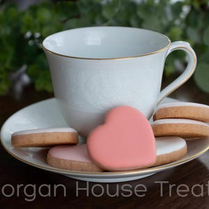 Six unwrapped iced cookies scattered and stacked in saucer with white china teacup. Greenery in background. Saucer and cup have gold accents. Morgan House Treats watermark at bottom of image.