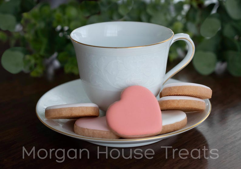 Six unwrapped iced cookies scattered and stacked in saucer with white china teacup. Greenery in background. Saucer and cup have gold accents. Morgan House Treats watermark at bottom of image.