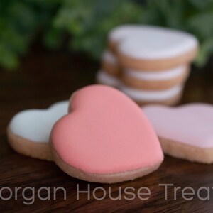 Close up stack of six unwrapped cookies, showing thickness and detail of pink cookie from side. Resting on dark wood table with greenery in background. Morgan House Treats watermark at bottom of image.