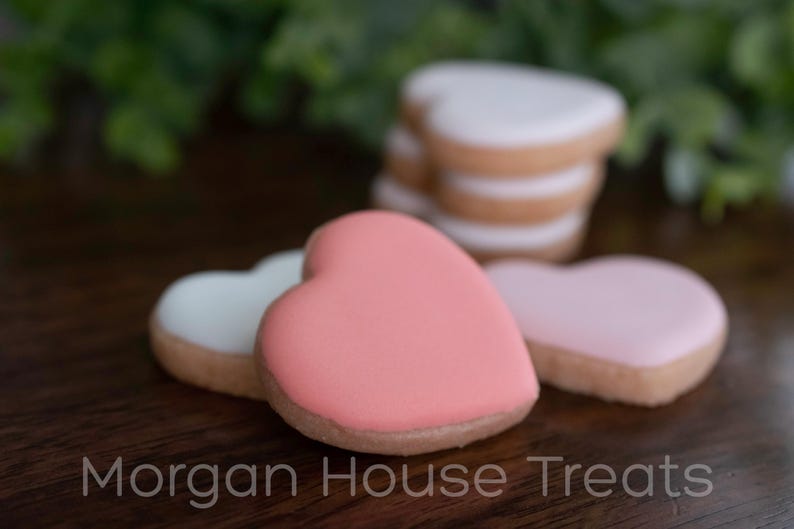 Close up stack of six unwrapped cookies, showing thickness and detail of pink cookie from side. Resting on dark wood table with greenery in background. Morgan House Treats watermark at bottom of image.