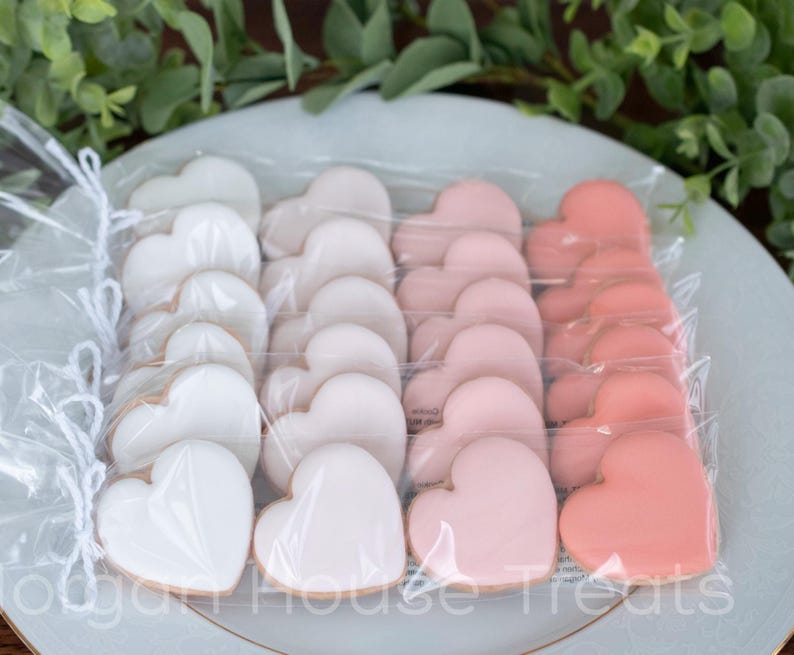 Six sleeves of pink mini heart cookies  on white china plate with greenery in background. Each sleeve holds four mini hearts simply decorated in ombre shades of royal icing, ranging from dark salmon to white. Sleeves tied with white baker's twine.