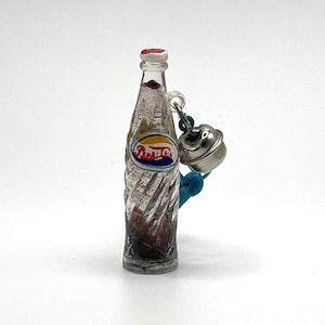 May include: A miniature glass Pepsi bottle charm with a silver bell and blue cord. The bottle has a clear, twisted design and a Pepsi logo. The bell is attached to the bottle with a small loop. The bottle is filled with a dark liquid.