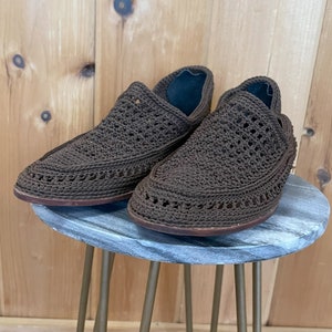 K&ouml;nnte beinhalten: Ein Paar braune geh&auml;kelte Slipper mit einem gewebten Muster. Die Schuhe stehen auf einem grauen Hocker mit Marmor-Muster und goldenen Metallbeinen.