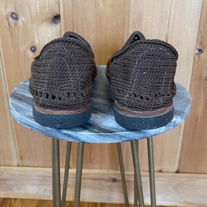 K&ouml;nnte beinhalten: Ein Paar braune geh&auml;kelte Schuhe mit Gummisohle, die auf einem Beistelltisch mit Marmor-Muster und goldenen Metallbeinen stehen.