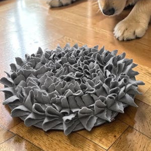 May include: A grey, circular pet bed with a ruffled design. The bed is made of a soft material and is designed to provide comfort for pets. The bed is on a hardwood floor.