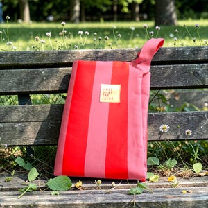 Può includere: Una borsa tote a righe rosse e rosa con manico, appoggiata su una panchina di legno. La borsa presenta un quadrato dorato con la scritta "MORE AMOR FOR FAVOR". Lo sfondo mostra un parco con alberi e fiori.