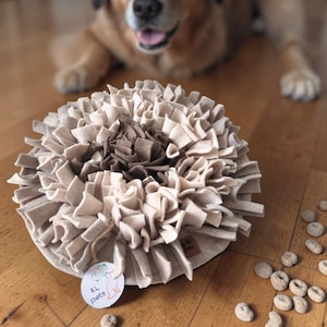 Op de afbeelding: Een ronde snuffelmat voor huisdieren gemaakt van gelaagde beige en donkerbruine viltstroken. De mat is voorzien van een rond wit label met de tekst KL pets en een kleine oranje lijntekening van een hond.