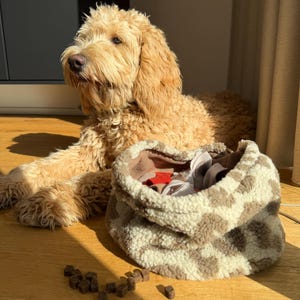 May include: A golden doodle dog lies on a wooden floor next to a soft, patterned dog bed. Scattered dog food pieces are in front of the bed. The dog bed is beige with brown spots and has a fluffy texture.